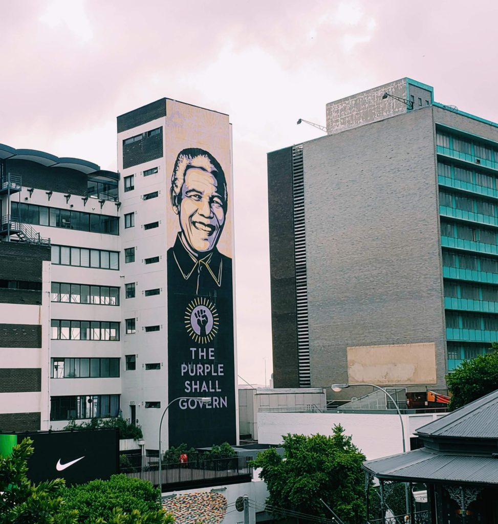 A photograph of an urban street in Johannesberg with a large mural of Nelson Mandela and the slogan 'The Purple Shall Govern.'