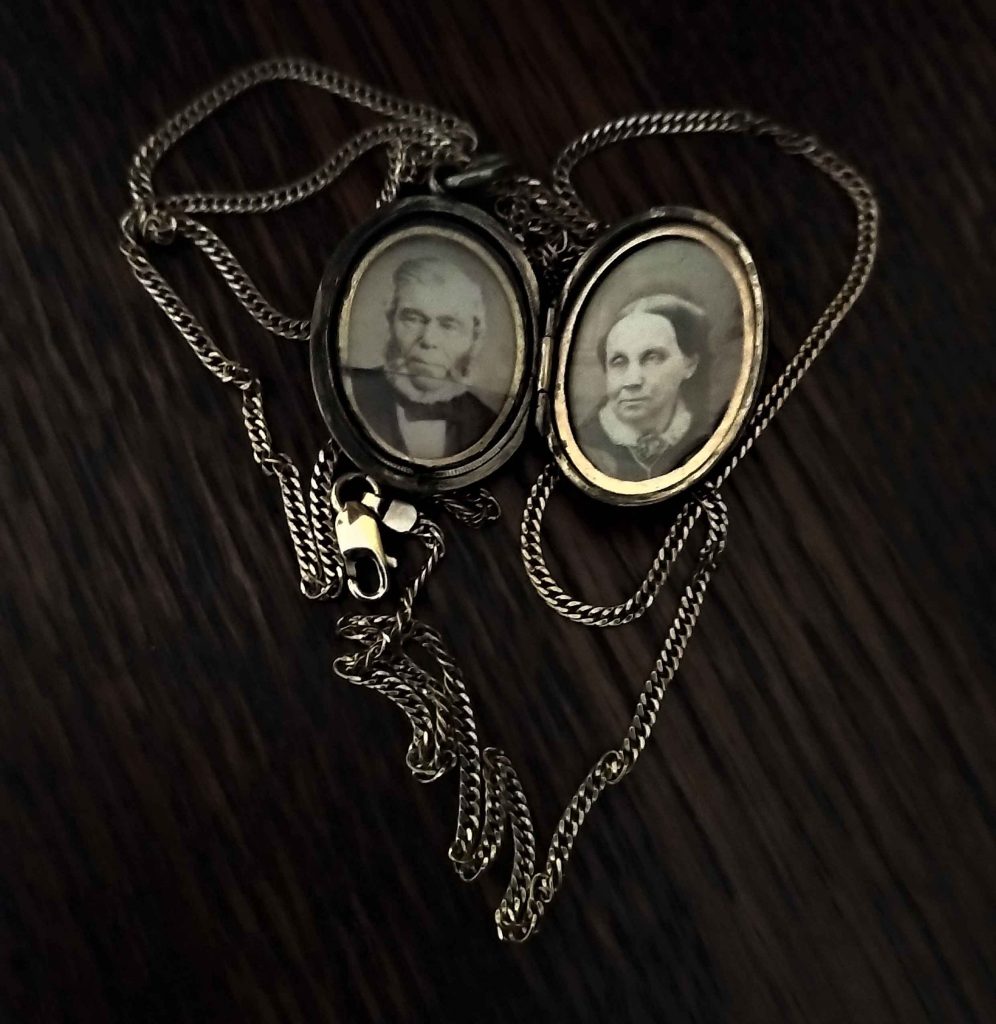 A silver locket attached to a silver chain on a wood table. Black and white photos on each side of the open locket. Man on right. Woman on left.