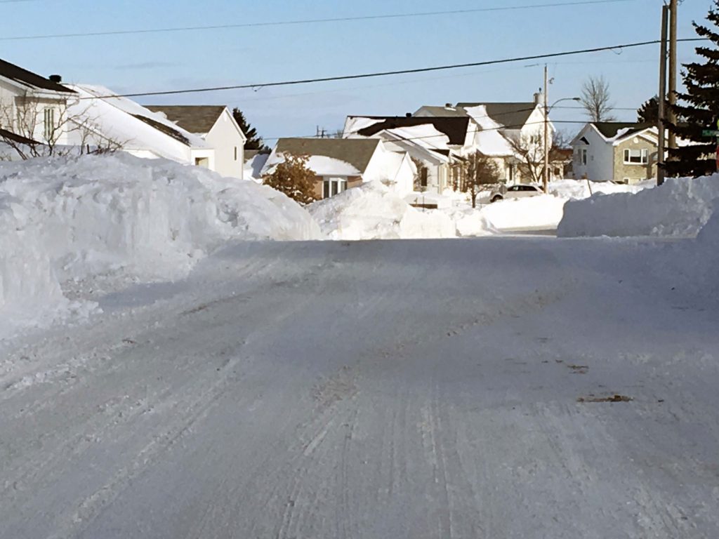 Vantage point from the middle of a residential street. Winter in Rimouski. High snowbanks line the street. Behind them are the tops of bungalows which vary in height.