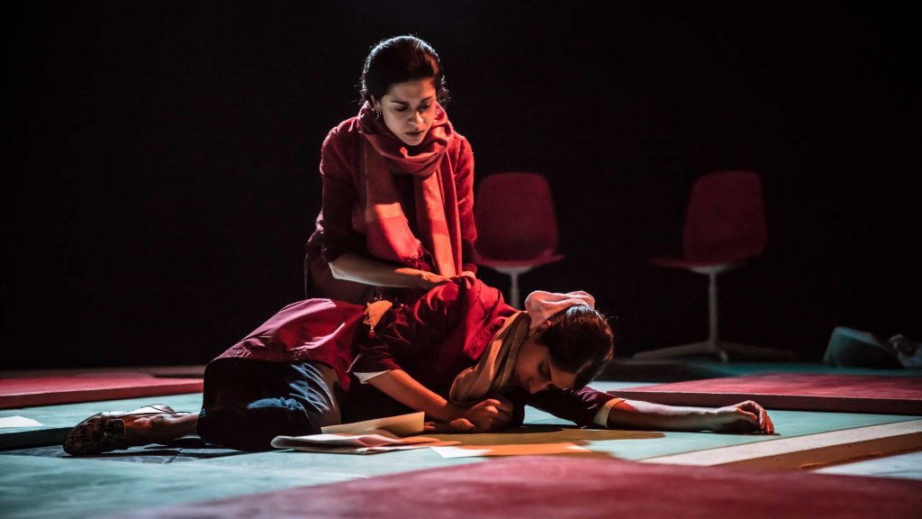 Two women at centre stage. The lighting is dramatic and dim but still clear. One woman lies on green and red-tiled flooring with her head down, in a partial fetal position, left arm outstretched. Sheets of paper are in front of her. A second woman crouches behind her with concern and right hand upon the first woman's shoulder.