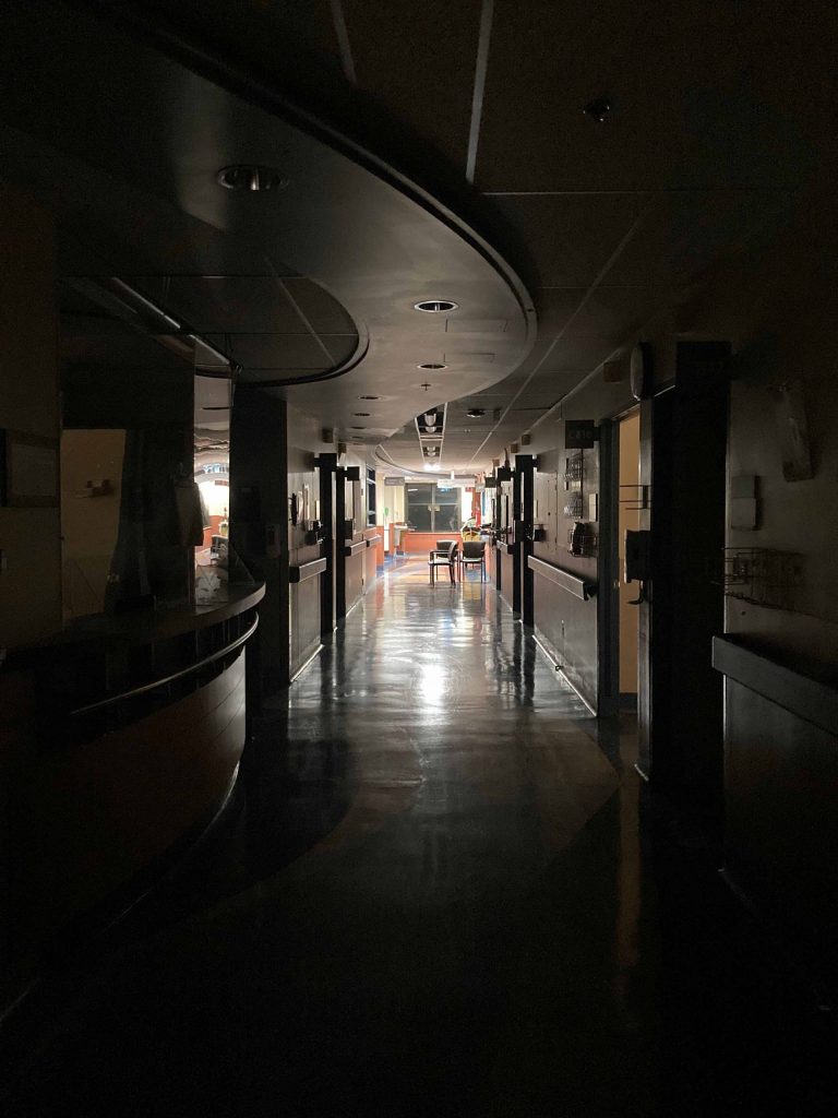 View of a hospital hallway. The lights are out in the foreground. To the left is a reception counter. The floors are shiny and reflect in the fluorescent light coming from a waiting room in the background at the other end of the hallway.