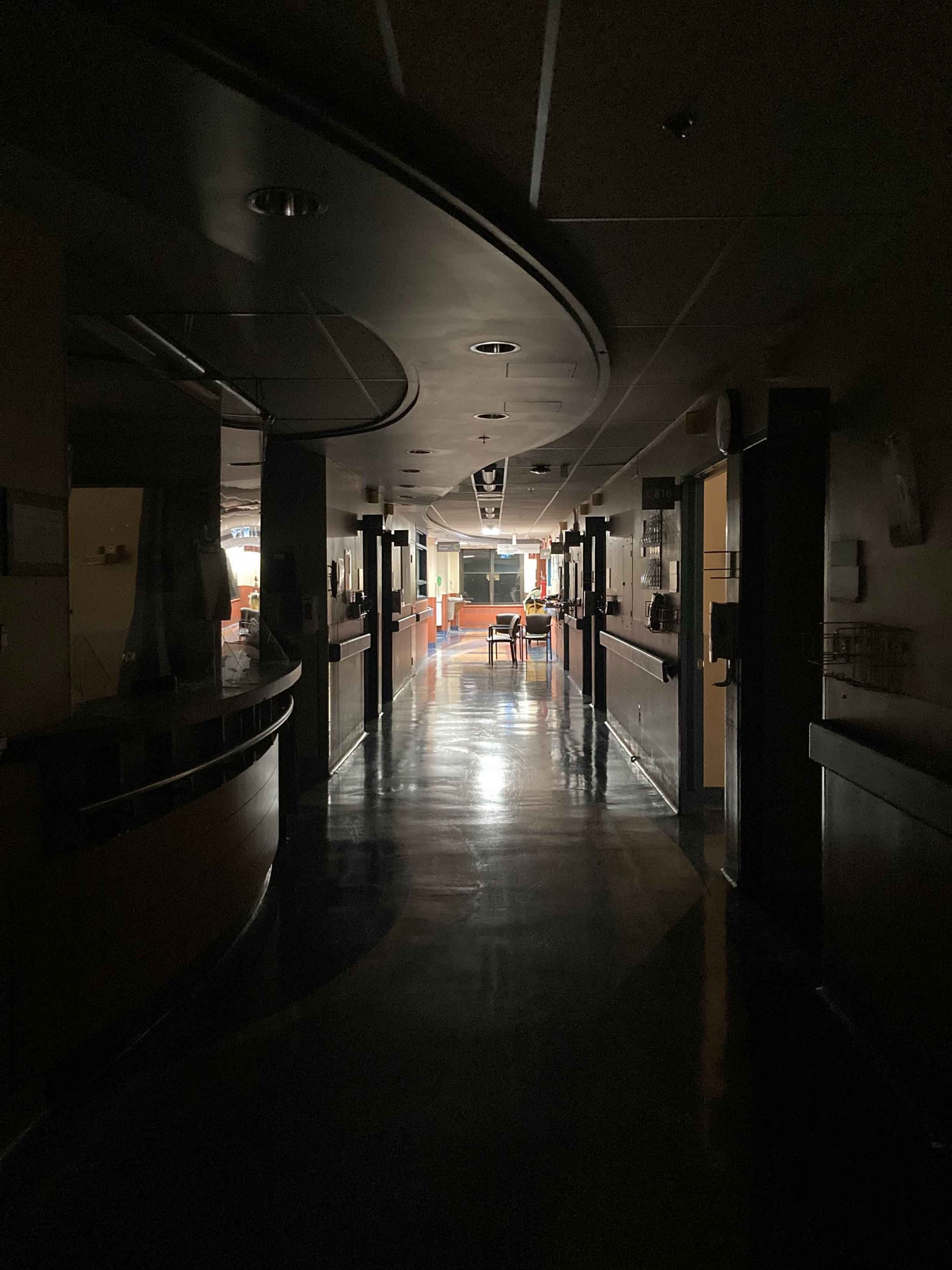 View of a hospital hallway. The lights are out in the foreground. To the left is a reception counter. The floors are shiny and reflect in the fluorescent light coming from a waiting room in the background at the other end of the hallway.