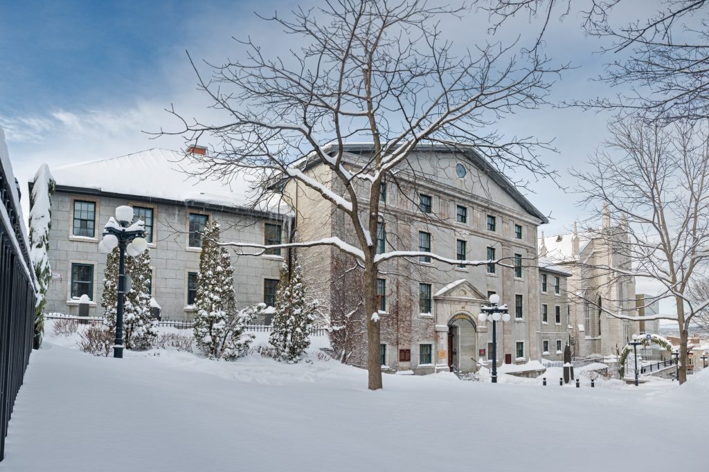 Horizontal photo of the Morrin Centre in Old Quebec City in winter. There's lots of snow in the foreground that covers the tree branches. The old large building sits in the mid-ground. It has lots of windows and a front door with large archway above it. 