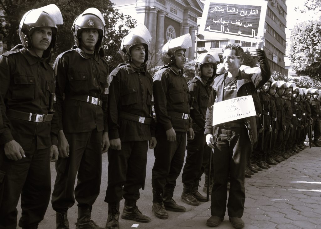 Black and white photo of military police. They stand side-by-side on a cobblestone street; helmet visors flipped up. A young man in casual clothes stands right before them. He smiles slightly, wears one placard with writing in English around his neck. He holds another with writing in Arabic in the air with his left hand. 