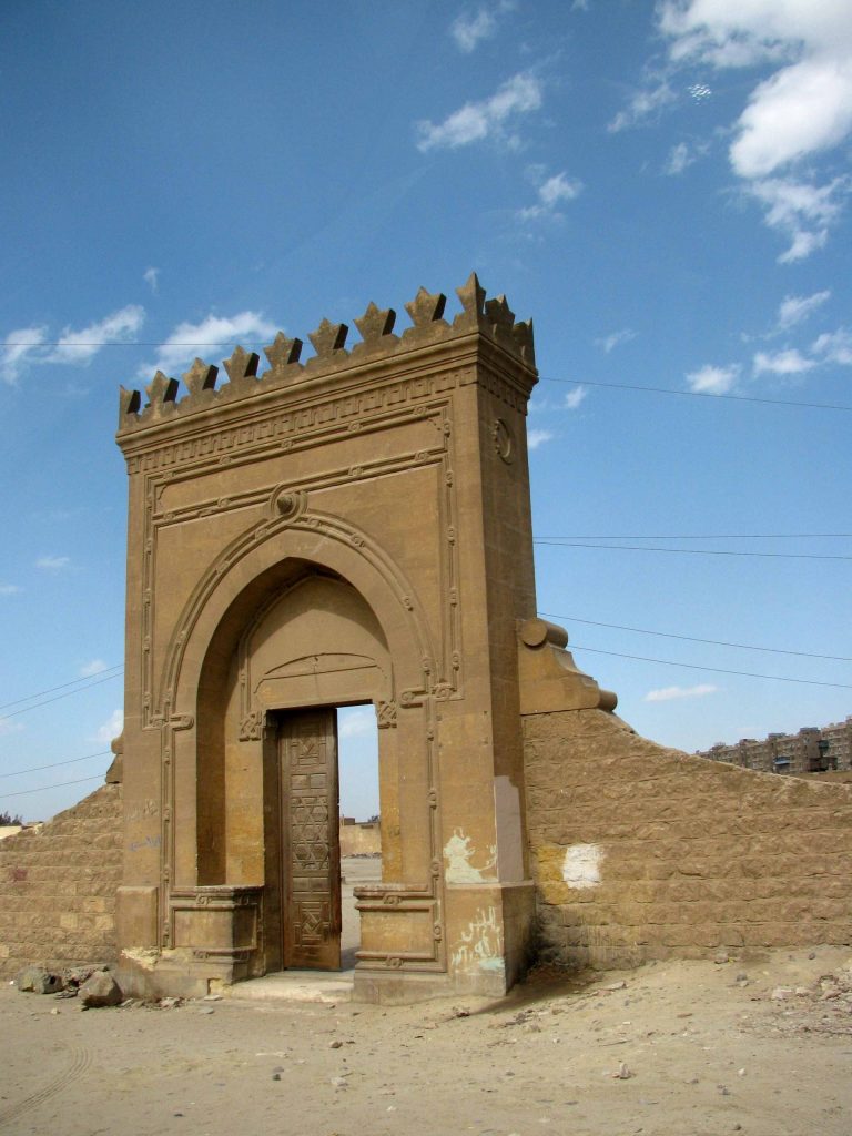 The entrance to an abandoned cemetery in Cairo. It is a large archway with a tall doorway built into it. One of the doors is closed; it is wood with shapes carved into it. The other half of the doorway is wide open. Beyond that and all around is clear blue sky and electricity power lines.