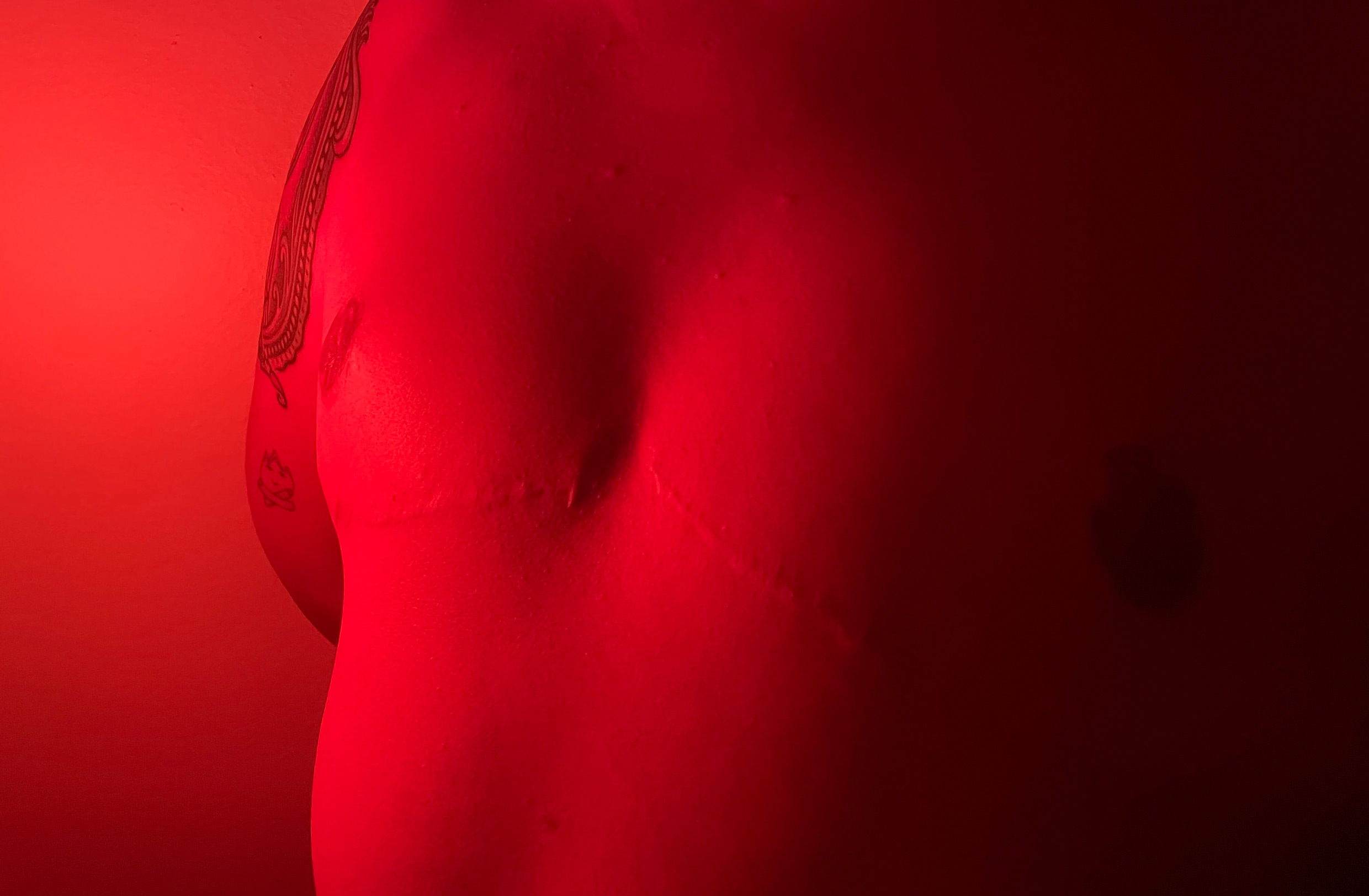 Photo of a non-binary person's chest after top surgery. There is a partial view of a tattoo on the person's body, which is set entirely within a filter of deep red light.
