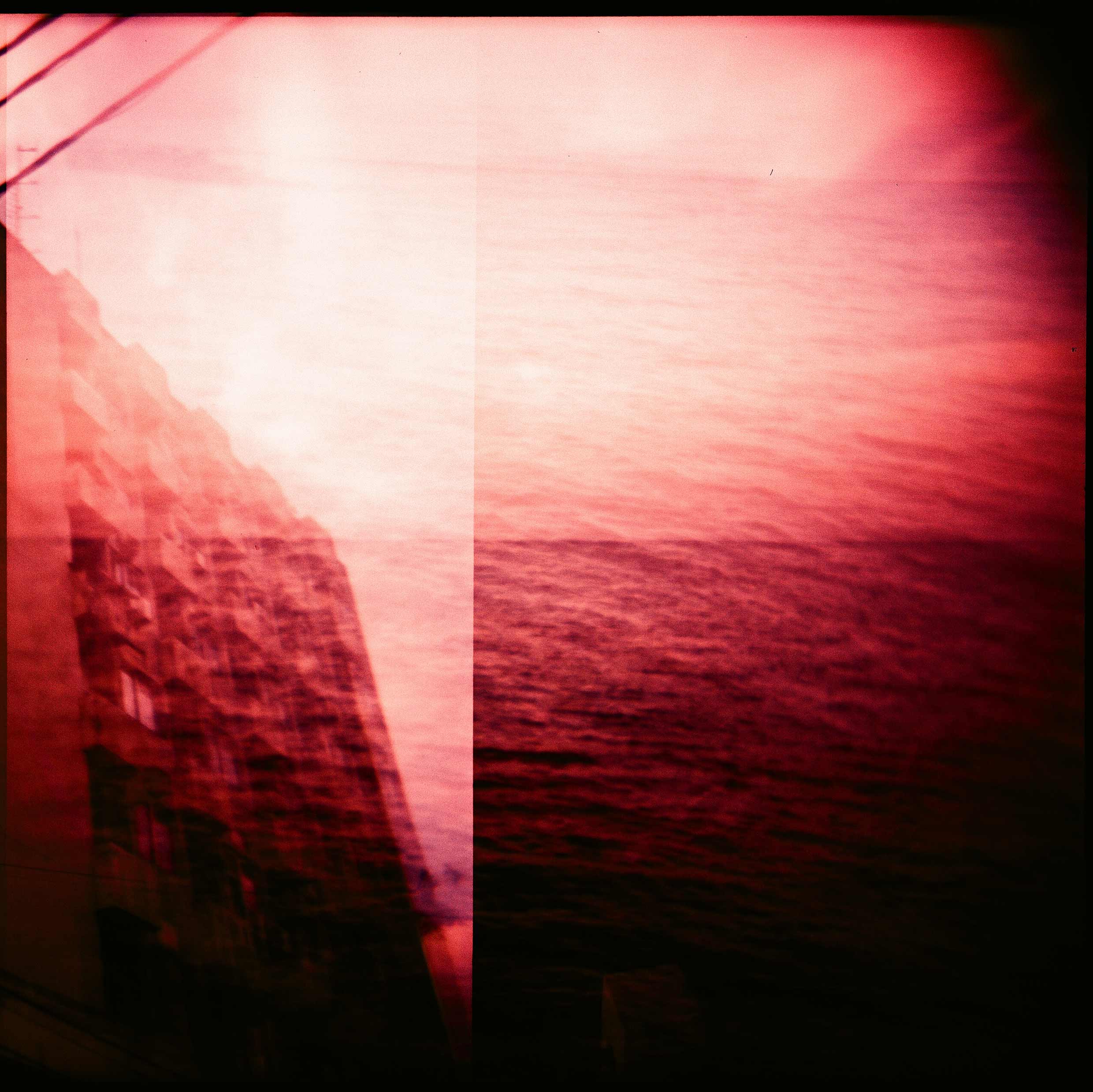 Photo framed by fish-eye lens tinted dark red with three semi-opaque layers; a large body of water and bright sky; a five or six-story concrete apartment building with balconies; three power lines. 