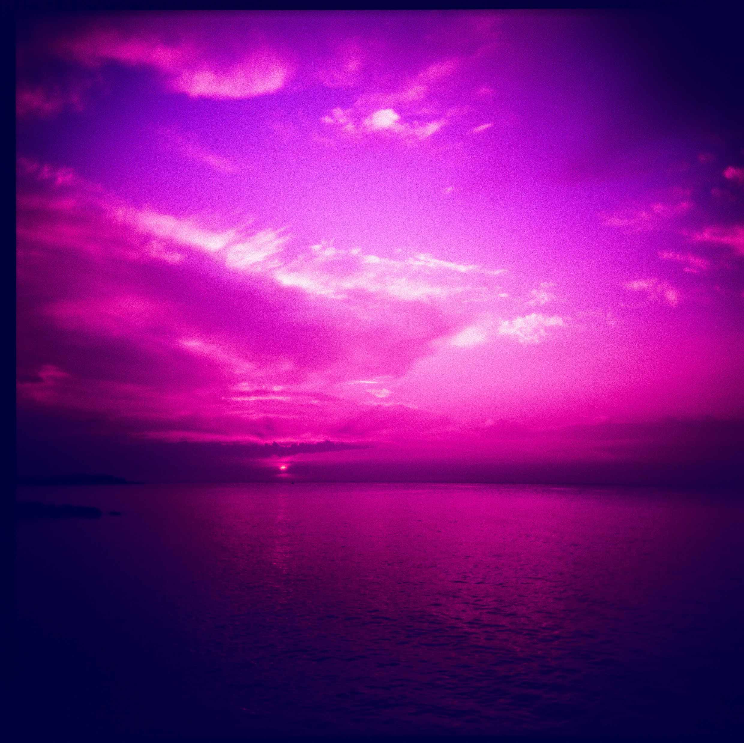 Photo framed by fish-eye lens tinted bright purple. A large body of water reaches into the horizon at sunset. Sunbeams reach into a partially-cloudy sky. 