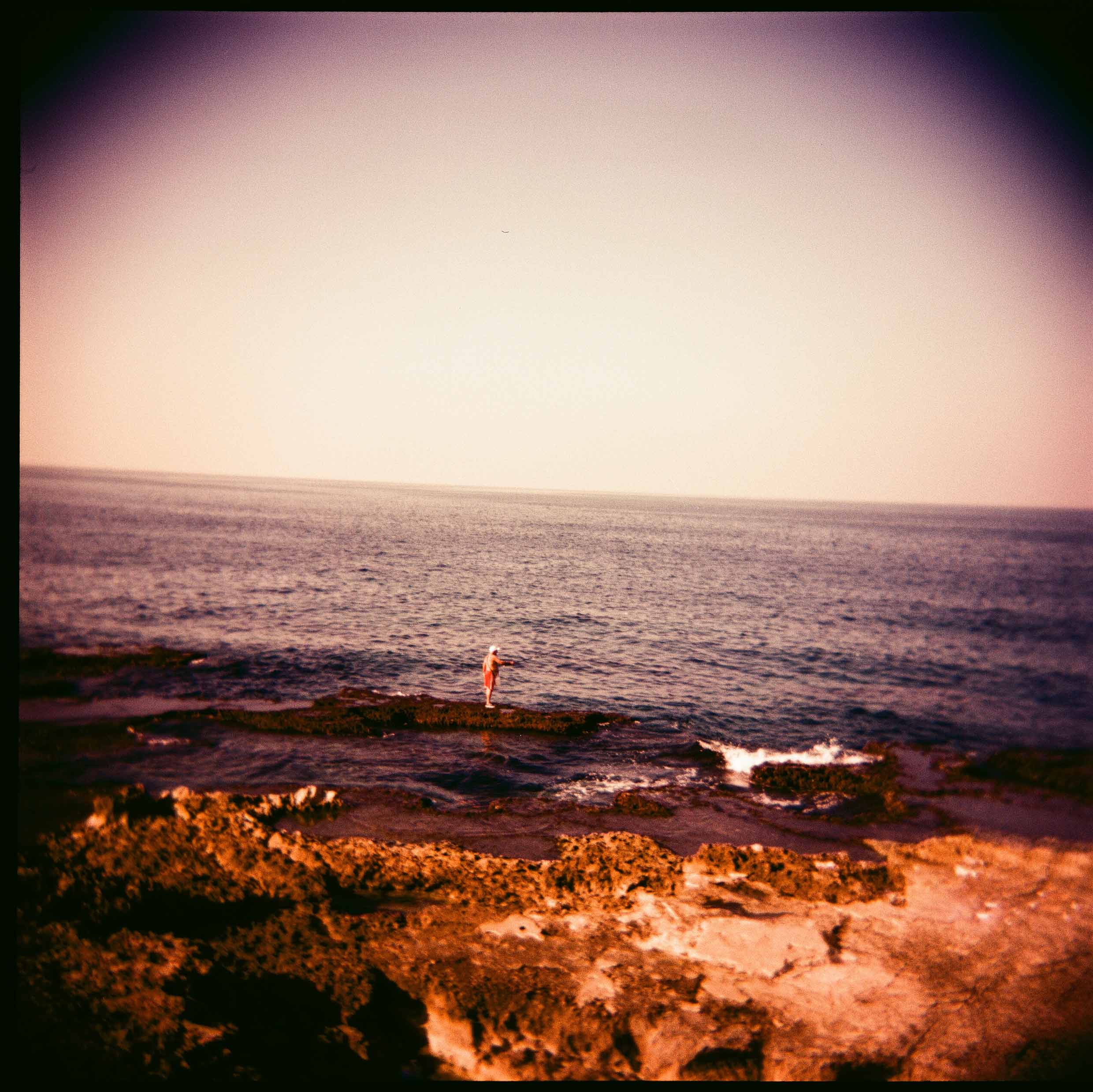 Photo tinted blue-yellow. Foreground: A rocky shore under a pale sky is covered with flora, then a narrow stretch of beach. A person wearing shorts, t shirt, and cap stands on a narrow sandbar; in the distance, the navy-blue water is calm. 