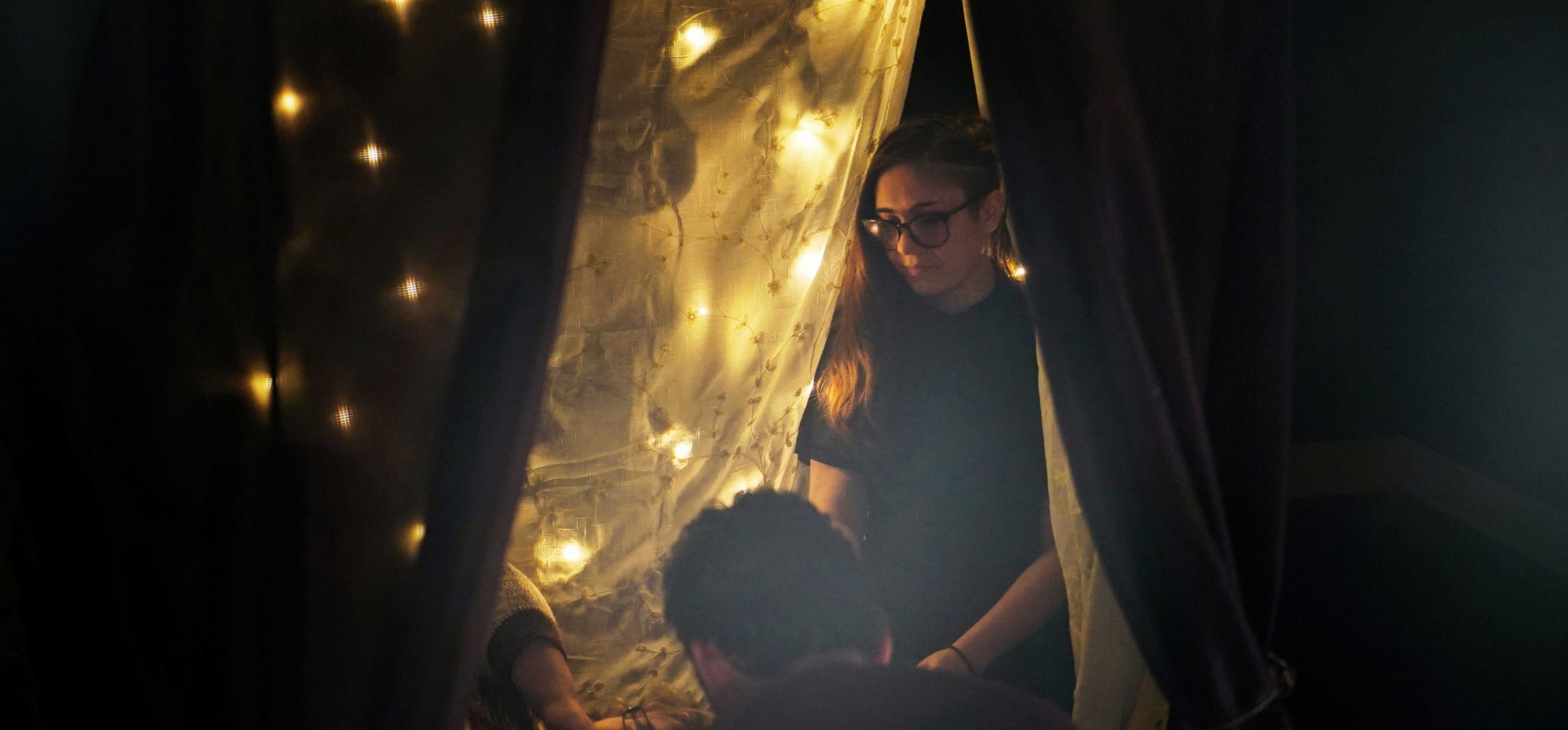 Three people sit on the floor facing each other. They're enclosed by curtains with string lights.