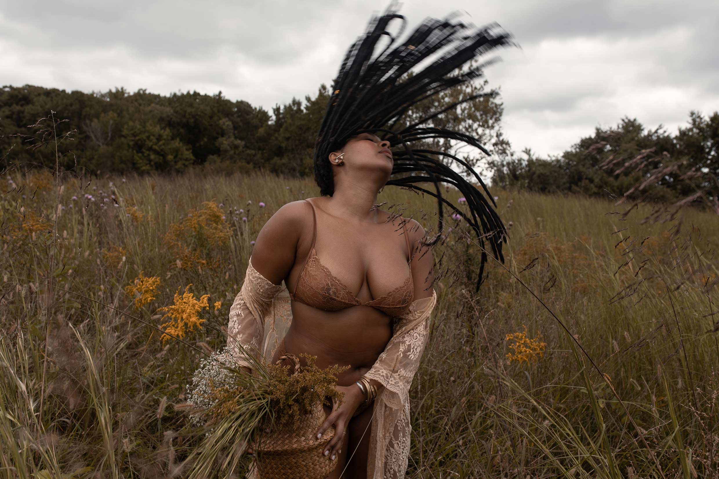 Curvy woman in grassland wearing beautiful undergarments holds a basket and is flipping her locs back.